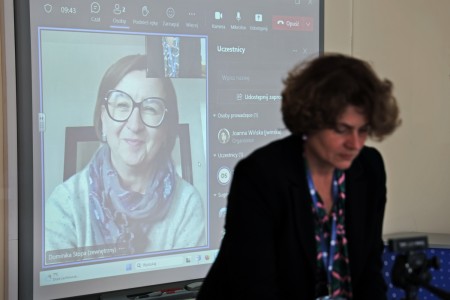 In the foreground of the photo, there is a woman - a conference participant - bent over documents. Behind her there is a multimedia screen showing a smiling woman during an internet remote call. Click to zoom the picture.