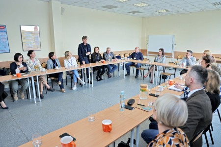 In the photo, a shot of the conference room, participants sitting at tables - men and women - looking towards a man standing and addressing the audience. Click to zoom the picture.