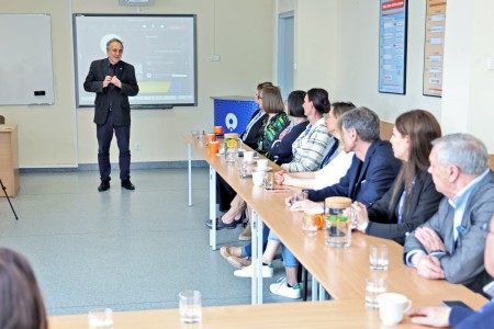 On the right, the photo shows participants seated at tables - men and women - looking toward a man standing in the middle of the room, addressing the audience. Click to zoom the picture.