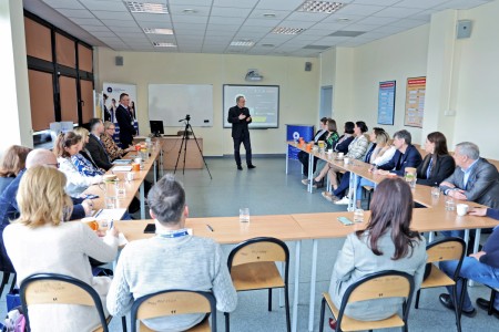 The photo shows a shot of the conference room from the back rows towards the front, participants are seated at tables, two men are standing addressing the audience. Click to zoom the picture.