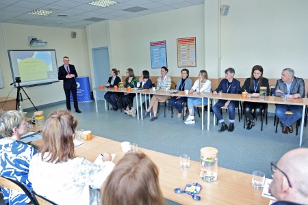 The photo shows a conference room, with participants-women and men- seated around tables. In the center there is a man standing and welcoming guests. Behind him there is a multimedia screen with the conference program displayed. Click to zoom the picture.