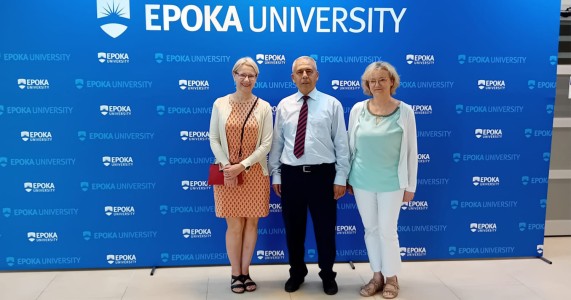 w środku Rektor Epoka University, prof.dr Ahmet Oztas; obok Rektora stoją wykładowczynie języka angielskiego – po lewej mgr Ewa Kowalska oraz po prawej stronie – mgr Dorota Hoffmann. Osoby te znajdują się  w gmachu Epoka University przy banerze promującym tę uczelnię. Kliknij, aby powiększyć zdjęcie.