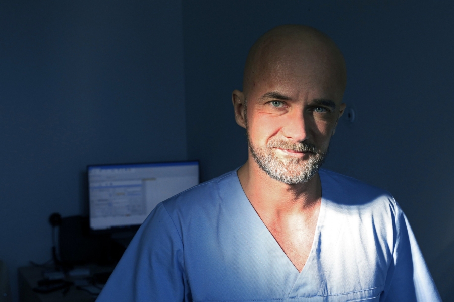 Portret bohatera wywiadu patrzącego na wprost, ubranego w fartuch weterynaryjny (jasnoniebieski). W tle włączony ekran komputera. Światło w pokoju przyciemnione.