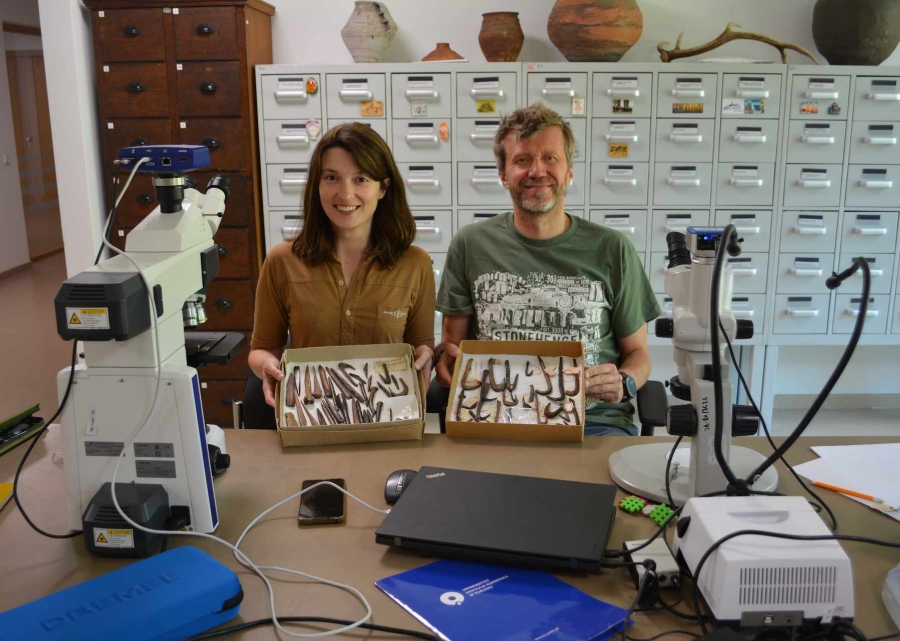 Naukowcy (kobieta i mężczyzna) trzymają w rękach pudełka z haczykami. Po prawej i lewej stronie stołu stoją mikroskopy. z tyłu za nimi znajdują się meble z dużą ilością szuflad